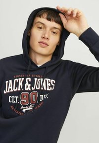 Felpa blu navy con cappuccio e cordoncino, con una stampa grafica bianca e marrone che dice "JACK & JONES 90" sul davanti. Tessuto morbido.