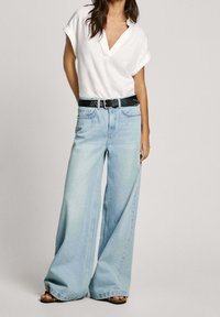 Vit kortärmad topp med V-ringning, ihop med ljusblå vida jeans och ett svart nitarbälte. Bruna öppna sandaler.