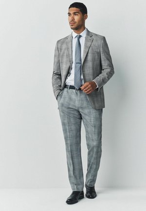 Grijze geruite suit met een getailleerde pasvorm, met een blazer met één knoop, bijpassende broek, wit shirt en een gestreepte stropdas. Zwarte nette schoenen.