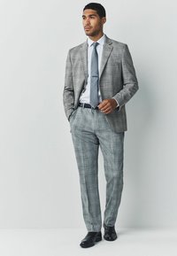 Traje de cuadros gris con un corte a medida, que incluye una americana de un solo botón, pantalones a juego, camisa blanca y una corbata estampada. Zapatos de vestir negros.