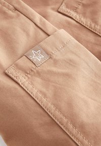 Beige stof med en glat tekstur, der har en syet lomme og et lille stjernesymbol broderet på kanten.