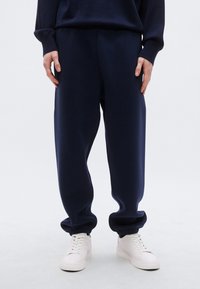 Μπλε σκούρο παντελόνι φόρμας με άνετη εφαρμογή, ριμπ μανσέτες και πλαϊνές τσέπες, συνδυασμένο με λευκά αθλητικά παπούτσια. Ομαλή υφή υφάσματος.