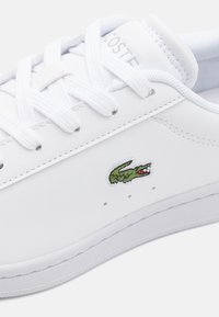 Vit lädersneaker med en slät yta, med en lågskaftad design, snörning och en liten grön krokodil-logotyp på sidan.