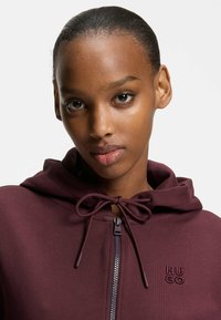 Felpa con cappuccio bordeaux con zip, cappuccio con coulisse, texture liscia e logo "HUGO" ricamato sul lato sinistro del petto.