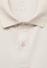 Camisa polo de color beige claro con una tela texturada, tapeta con tres botones y cuello puntiagudo. Presenta un acabado sutil y suave.