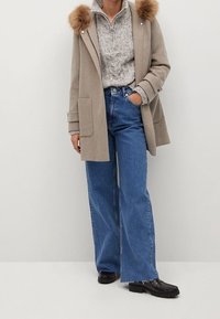 Abrigo de lana beige con cuello de piel, suéter de punto gris debajo y jeans de pierna ancha azul de cintura alta. Zapatos chunky negros completan el atuendo.