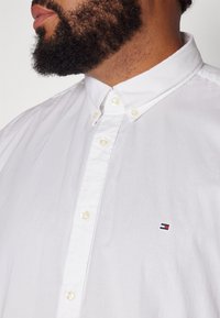 Uomo con la barba che indossa una camicia bianca con bottoni, caratterizzata da un piccolo logo ricamato rosso, bianco e blu sul petto.