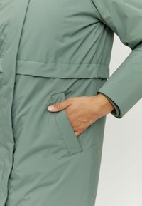 Green, padded coat with a belt, featuring a vertical seam and side pocket. Smooth texture and tailored fit with a cuff detail.