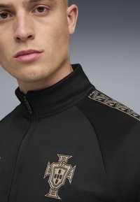 Κοντινό πλάνο ενός ατόμου που φοράει μαύρο μπουφάν με φερμουάρ και μπεζ έμβλημα στο στήθος, καθώς και διακοσμητική ρίγα στον ώμο.