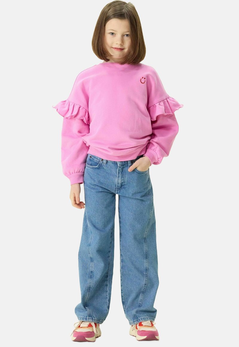 Sweatshirt rose avec des manches à volants, logo smiley, associé à un jean baggy bleu clair et des baskets multicolores.
