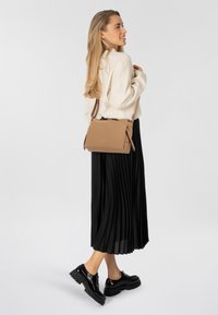 Tasche aus braunem Leder im Querformat mit glatter Textur und Reißverschluss. Ein schwarzer plissierter Rock und glänzende schwarze Schuhe vervollständigen das Outfit.