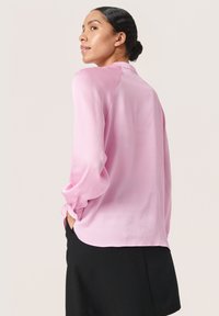 Rosa langærmet bluse i tekstureret stof med krave og knapmanchetter, parret med sorte bukser. Kun set fra ryggen.
