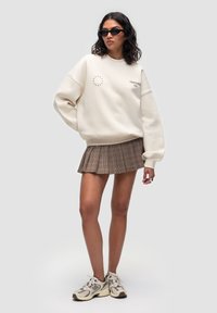 Felpa oversize color crema con stampa di stelle, abbinata a una gonna a pieghe a quadri marrone. La modella indossa occhiali da sole neri e scarpe da ginnastica beige.