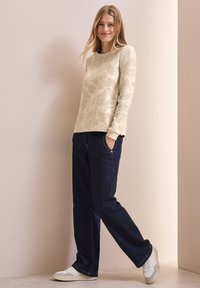 Cremfarbener Pullover mit Blattmuster, kombiniert mit dunkelblauen, geraden Jeans und weißen Sneakers, vor einem neutralen Hintergrund.