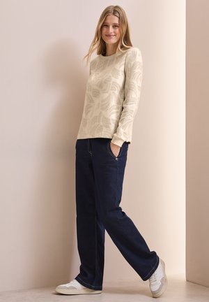 Suéter crema con patrón de hojas, combinado con jeans de pierna recta azul oscuro y zapatillas blancas, sobre un fondo neutral.