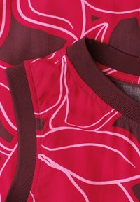 Gros plan sur un tissu rouge avec des lignes abstraites roses et une garniture bordeaux foncé au col et à l’emmanchure, montrant une partie d’un vêtement sans manches.