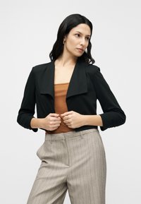 Μαύρο κοντό σακάκι με φαρδύ γιακά και μανίκια γυρισμένα, σε συνδυασμό με καφέ τοπ και ανοιχτό γκρι παντελόνι με λεπτές ρίγες.