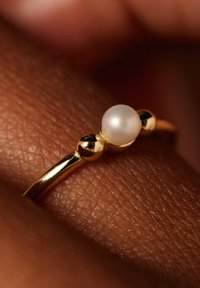 Gouden ring met een gladde band, een centrale witte parel en twee kleine sferische gouden accenten aan weerszijden. Textuur van de huid op de achtergrond.