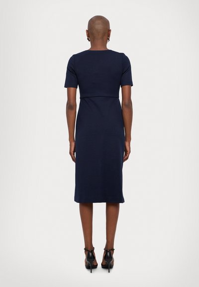 Robe midi marine à manches courtes, corsage ajusté et jupe évasée. Texture de tissu lisse, design minimal, portée avec des chaussures à talons hauts noires.