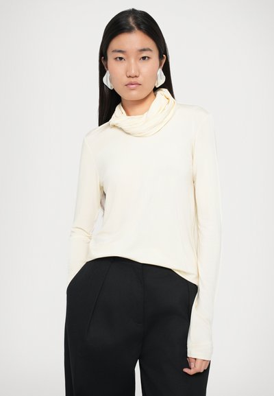 Crème lange mouw top met een colkraag, gemaakt van soepel materiaal, gecombineerd met zwarte hoge taille broeken. Model draagt grote witte oorringen.