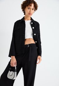 Chaqueta negra texturizada con bolsillos frontales y botones en forma de flor, combinada con una blusa corta blanca y pantalones negros de tiro alto, sosteniendo un bolso pequeño.