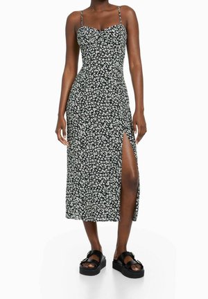 Kvinde iført sort og hvid blomstret midikjole med spaghetti-stropper og slids op til låret, kombineret med sorte platform-sandaler.