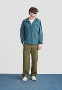 Νεαρός άνδρας που φοράει τζάκετ Columbia σε τιρκουάζ χρώμα, ελαι πράσινο παντελόνι cargo, λευκά αθλητικά παπούτσια και μαύρα γυαλιά ηλίου, στέκεται σε μπλε χαλί κοιτάζοντας πλάγια.