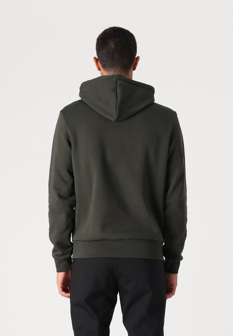 Haut à capuche vert foncé en tissu doux, doté d'une capuche standard et de poignets côtelés. Coutures visibles et coupe décontractée.