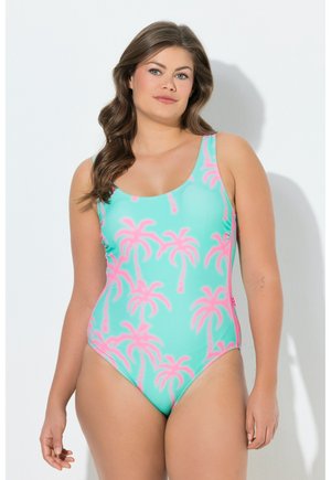 Kobieta z długimi falowanymi włosami, ubraną w turkusowy strój kąpielowy z różowymi wzorami palm, stoi przy białej ścianie.