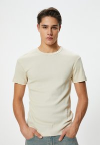Beige ribbet t-shirt med korte ærmer og rund hals, med en slim fit og glat tekstur. Båret med hænderne i lommerne.