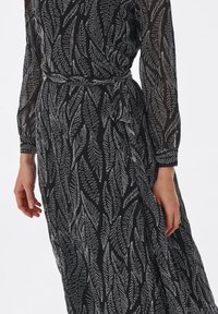 Vestido negro con un estampado de hojas en blanco. La tela es transparente, con mangas largas y un detalle de cintura ajustada, que realza su silueta.