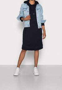 Jeansjacka i ljusblått över en svart huvad klänning. Vita sneakers med subtila detaljer. Slät tygtextur och avslappnad passform.