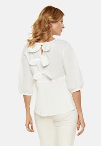 Blusa bianca con maniche a sbuffo, caratterizzata da un design con apertura a chiave sul retro e tre fiocchi decorativi. Tessuto morbido, vestibilità rilassata.