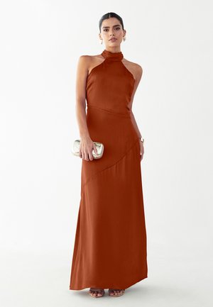 Kobieta w długiej sukni na szyję w kolorze rdzawego brązu, trzyma małą metaliczną kopertówkę, z złotymi kolczykami kołami i zegarkiem.