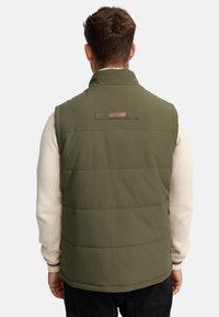Vesta de puf verde olive cu guler înalt, cusături verticale și o etichetă cu patch la spate. Purtați peste un pulover de culoare crem.