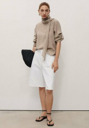 Model wearing a beige sweater with a tied neck, white shorts, and black strappy sandals, holding a round black woven bag against a neutral backdrop.