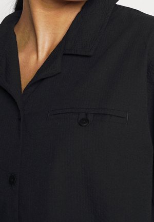 Camisa negra abotonada con tela texturizada, que incluye cuello y un bolsillo en el pecho con cierre de botón.