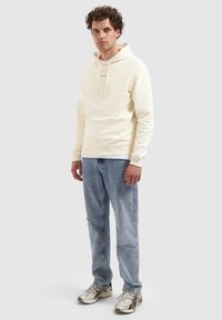 Jonge man met krullend haar, gekleed in een crèmekleurige hoodie met de tekst "PURE PATH", lichtblauwe jeans en zilveren hardloopschoenen, staande op een grijze achtergrond.