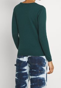 Dunkelgrüner Pullover mit langen Ärmeln, strukturiertem Rautenmuster und geripptem Saum, kombiniert mit blauen Batik-Cargohosen.