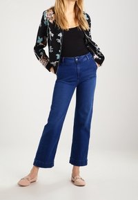 Veste bomber noire à motif floral avec des accents bleus et roses, associée à un jean large taille haute bleu et à des chaussures légères roses enfilables.