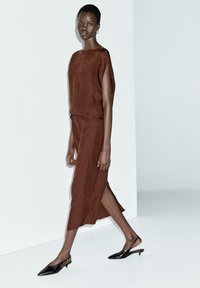 Hnědé lněné šaty s volným střihem a bočními rozparky, spárované s černými lodičkami s špičatou špičkou a otevřenou patou. Minimalistický design na jednobarevném pozadí.