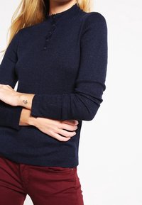 Granatowy sweter z prążkowanego dzianiny z wysokim kołnierzem, detalami z guzikami przy szyi i długimi rękawami, zestawiony z bordowymi spodniami.