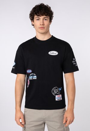 Junger Mann mit lockigem Haar, der ein schwarzes T-Shirt mit mehreren weißen und blauen Aufnähern und beige Cargoshorts vor einem einfarbigen Hintergrund trägt.
