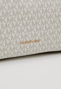 Beżowa torebka wykonana z fakturowanej tkaniny z powtarzającym się wzorem logo "MK", ozdobiona złotą tabliczką z napisem "MICHAEL KORS".