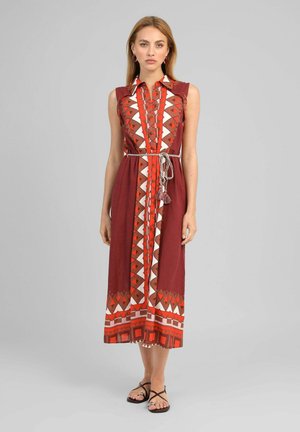 Femme portant une robe rouge sans manches, longueur genou, avec des motifs géométriques, une fermeture à boutons sur le devant et une ceinture tressée blanche, associée à des sandales noires.
