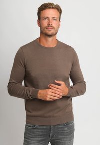 Bruine gebreide trui met een ronde halslijn en lange mouwen, voorzien van geribde mouwen en zoom. Gedragen met grijze jeans, eenvoudig ontwerp zonder patronen.