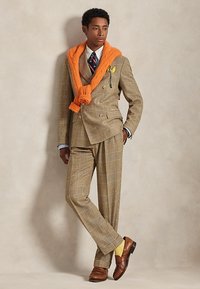 Άνδρας με καφέ καρό κοστούμι, μπλε γραβάτα, κίτρινο μαντήλι στο πέτο, πορτοκαλί πουλόβερ περασμένο πάνω από τους ώμους, κίτρινες κάλτσες και καφέ μοκασίνια.