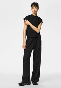 Mlada osoba s kratkom kosom u crnoj majici bez rukava, širokim hlačama s prugama i crnim tenisicama, stoji na bijeloj pozadini.