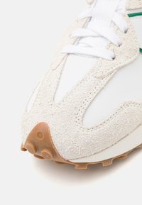 Gros plan sur la pointe d'une chaussure de sport en daim beige et maille blanche, avec des lacets blancs et des picots en caoutchouc marron sur une semelle, sur fond blanc.
