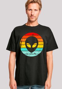Schwarzes T-Shirt mit einem verwitterten Grafikdesign eines Alienkopfes und konzentrischen Streifen in Rot, Orange und Türkis. Lässiger Schnitt, kurze Ärmel.